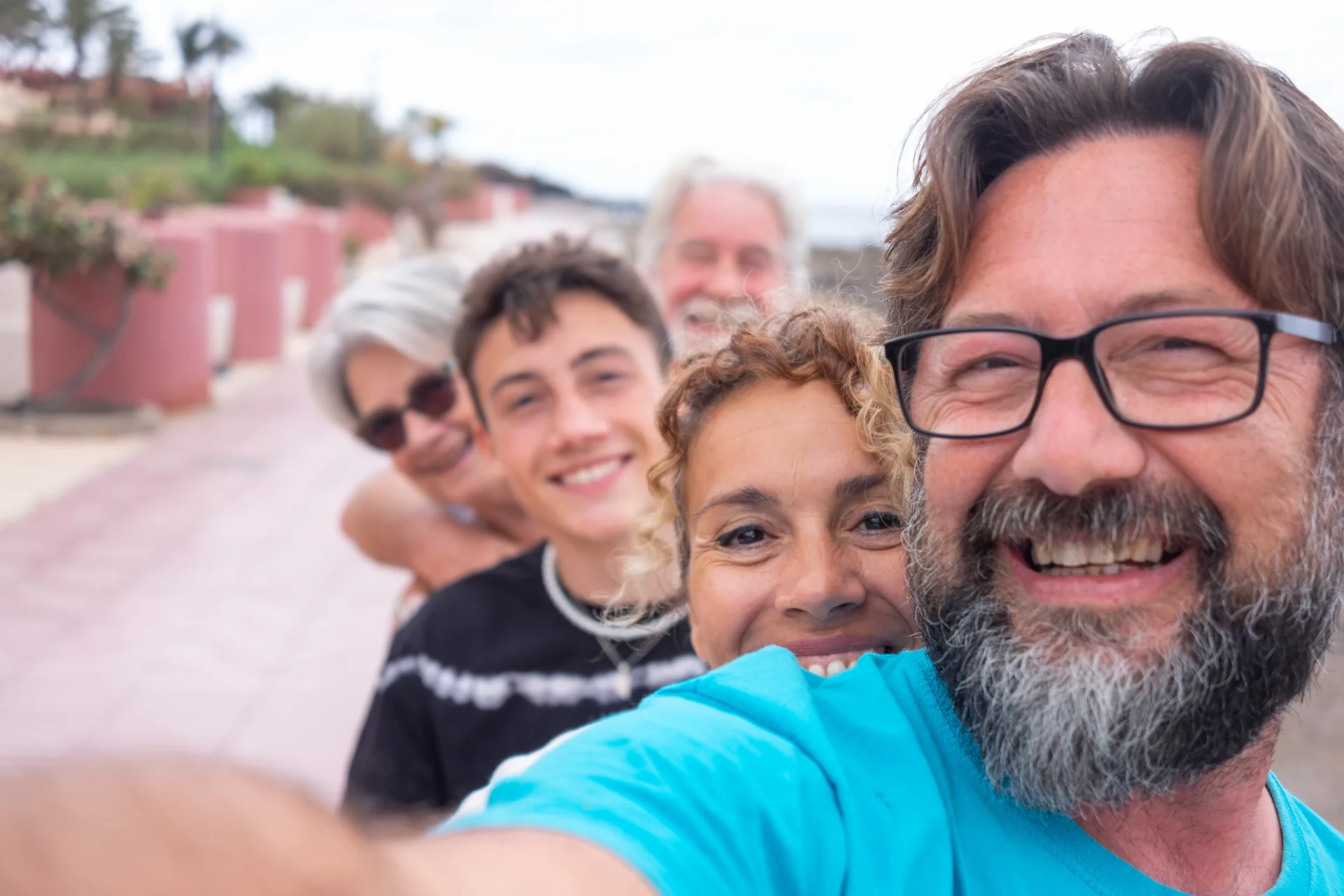 A multi-generational family taking a selfie, with a middle-aged man in the foreground smiling wide. the background is slightly blurred.