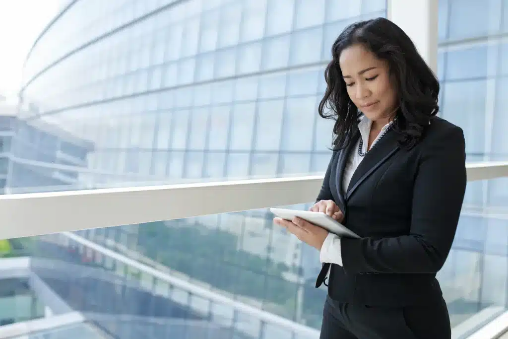 Businesswoman using a tablet computer on a balcony with a modern office building in the background.
