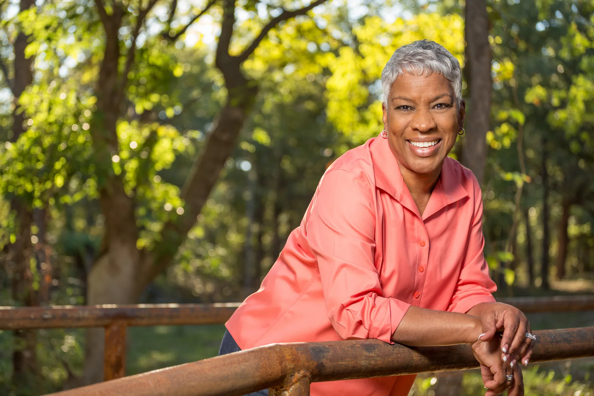 An older african american woman with short gray hair, smiling in a pink shirt, leans on a fence in a sunlit park.