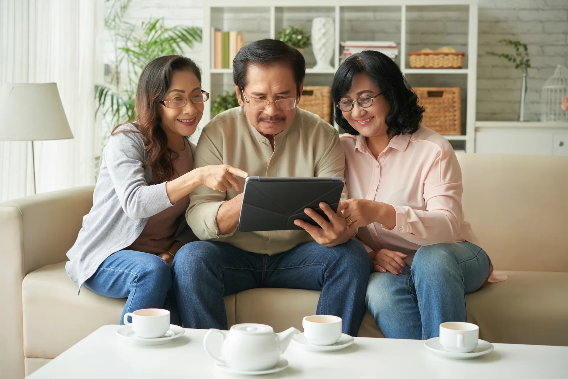Three elderly people sitting on a sofa, looking at a tablet and smiling, with cups of tea on the table in front of them.