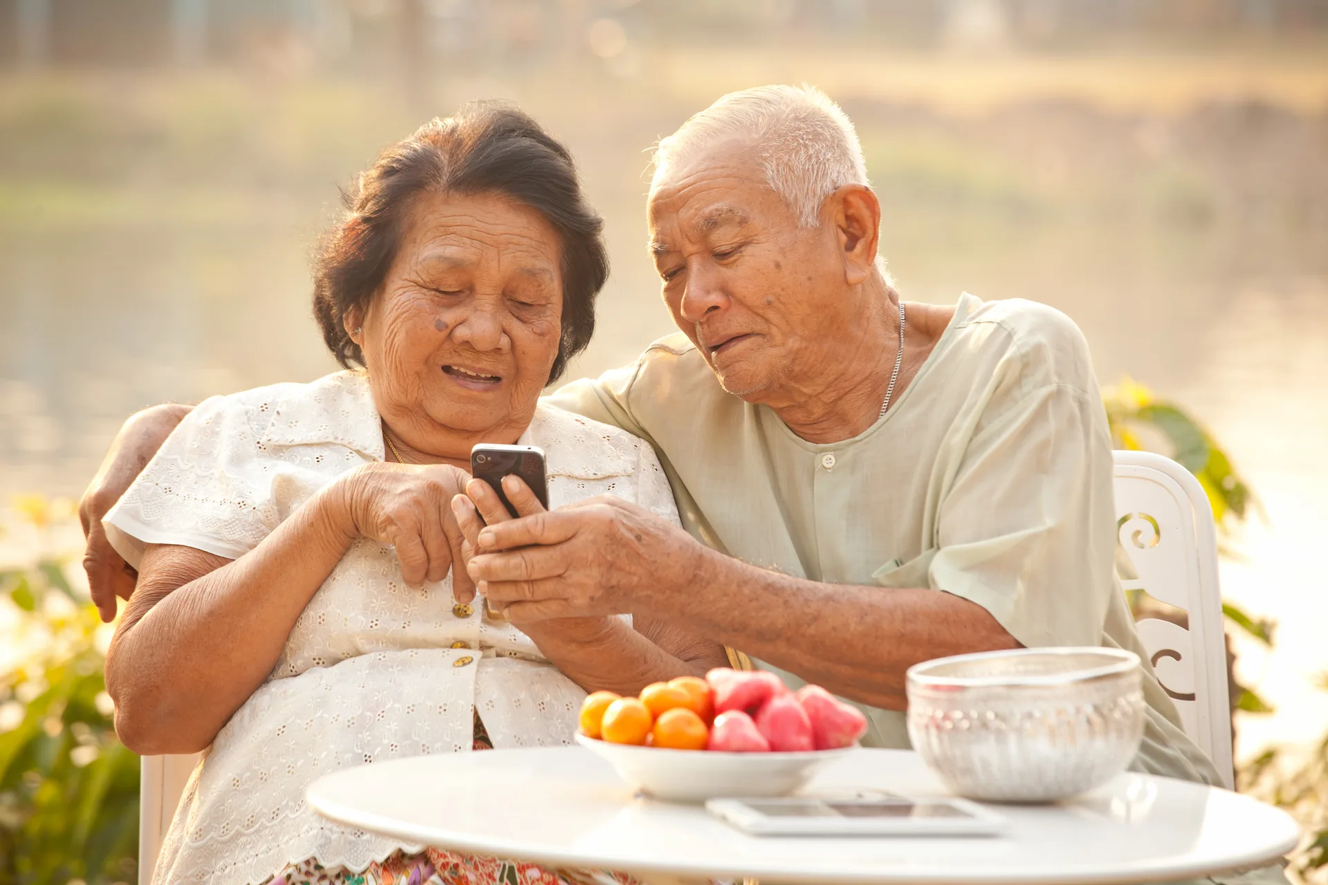 An elderly couple looks at a smartphone together while sitting at an outdoor table with fruit.
