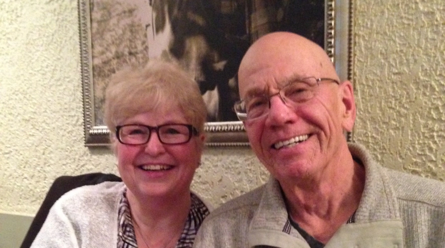 A smiling elderly couple with glasses, sitting together in front of a framed black and white photograph on a wall, exemplifying the warmth and support in dementia care.