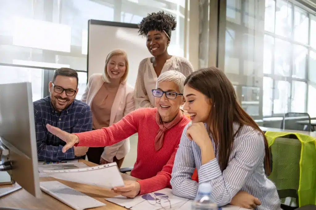 Diverse group of five colleagues discussing work around a computer in a bright office space.
