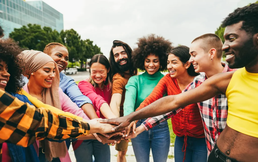 A diverse group of young adults smiling and stacking hands together in a show of unity outdoors.