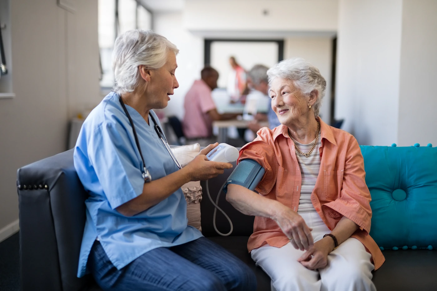 A nurse checking the blood pressure of an elderly woman in a bright room, both smiling and engaging in conversation.
