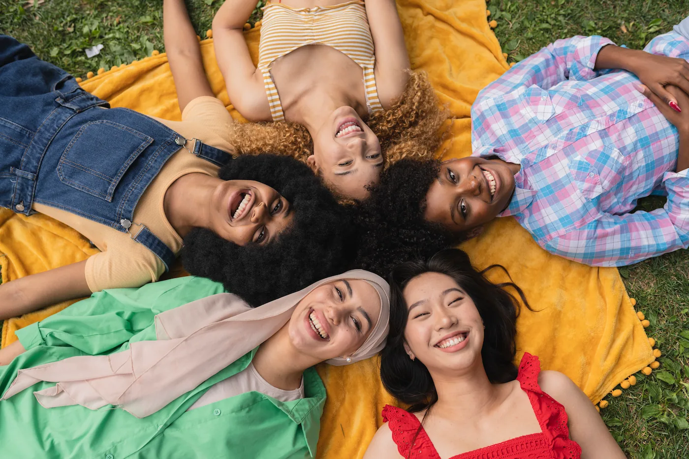 Five diverse young women lying head to head on a yellow blanket in a park, smiling and enjoying each other's company.