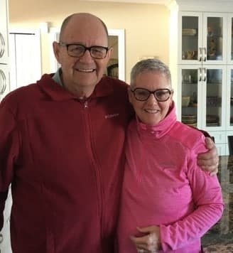 An elderly couple, Gaylene and her partner, smiling, wearing matching pink and red jackets, standing arm-in-arm inside a home with white doors in the background.